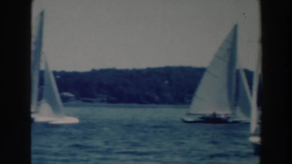1964 Tustin, Wisconsin home-movie frame showing several small sailboats on open water with a tree-lined shore behind them.