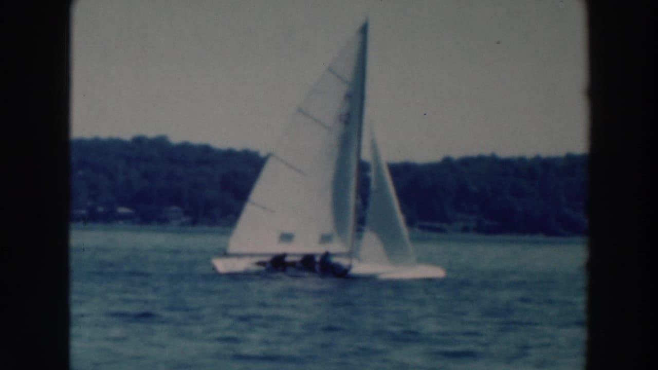 1964 Tustin, Wisconsin frame with sailboats crossing a lake, crews visible under white sails and wooded shoreline beyond.