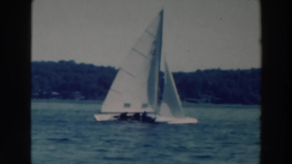 1964 Tustin, Wisconsin frame with sailboats crossing a lake, crews visible under white sails and wooded shoreline beyond.