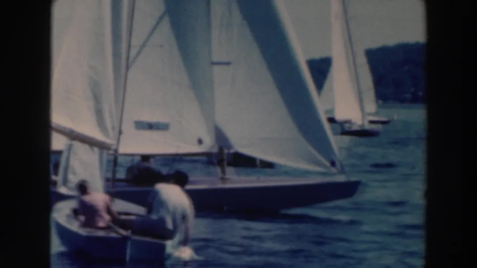 1964 Tustin, Wisconsin frame with a close view of people in a small boat beside a sailboat with raised white sails.