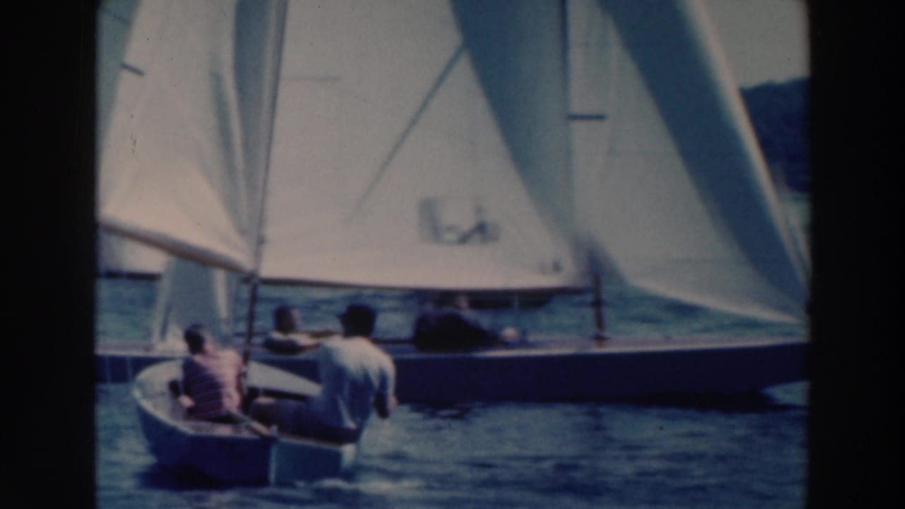 Small sailboat with two people near larger white-sailed boats on a lake.