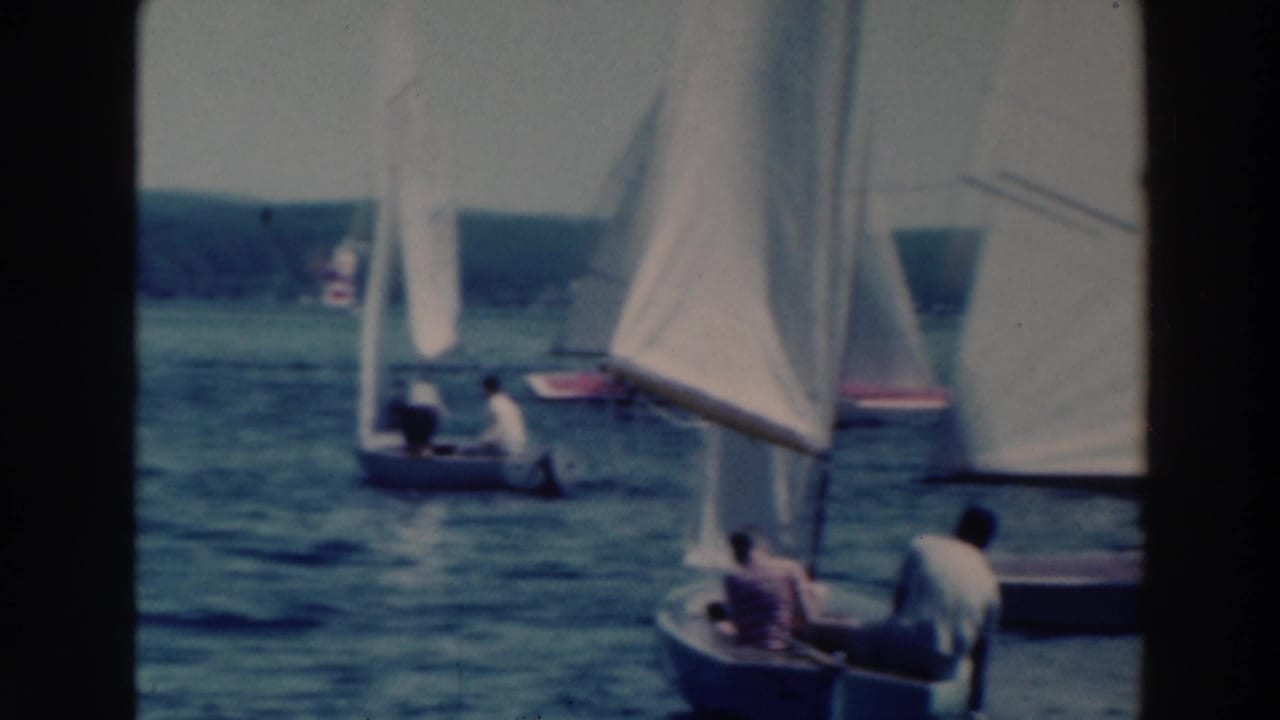 Several white-sailed boats move across lake water with a small boat in the foreground.