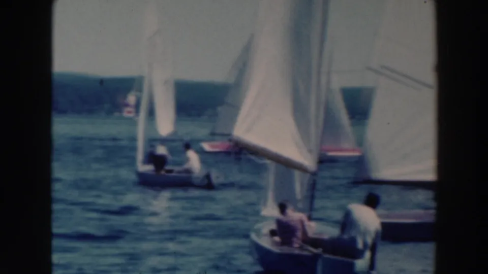 A 1964 home-movie frame from Tustin, Wisconsin, showing sailboats clustered on a lake with two people in a foreground dinghy.
