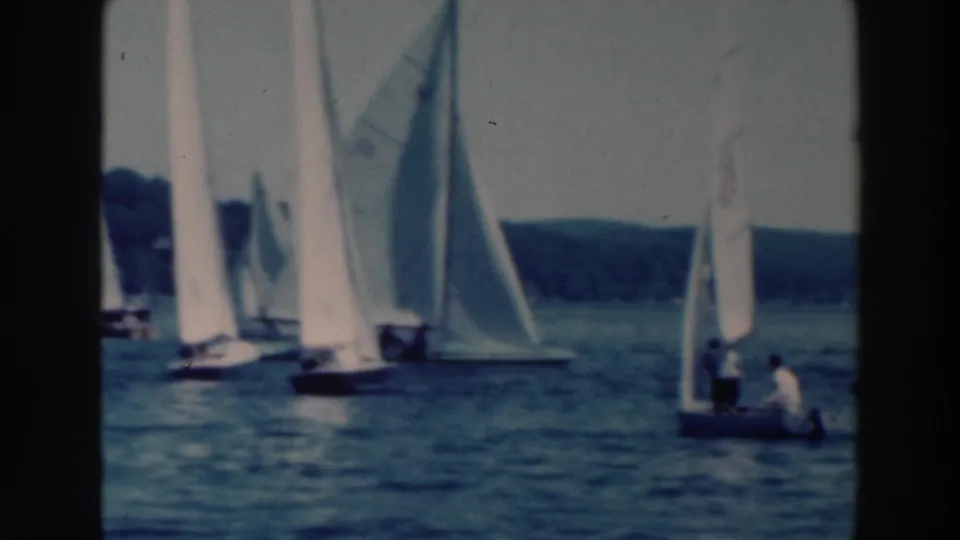Soft 8mm frame showing white sails and small boats on a Wisconsin lake, with distant trees and seated sailors visible.