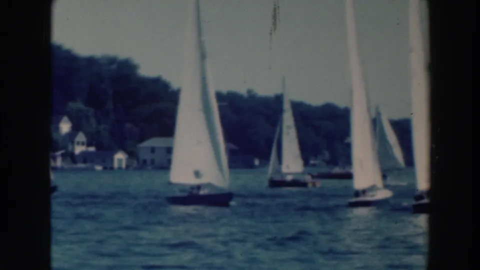 1964 Wisconsin 8mm frame showing several sailboats crossing a lake near a wooded shore and distant houses.