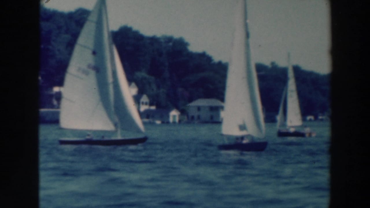 Sailboats with white sails pass near a wooded lakeshore and small buildings.