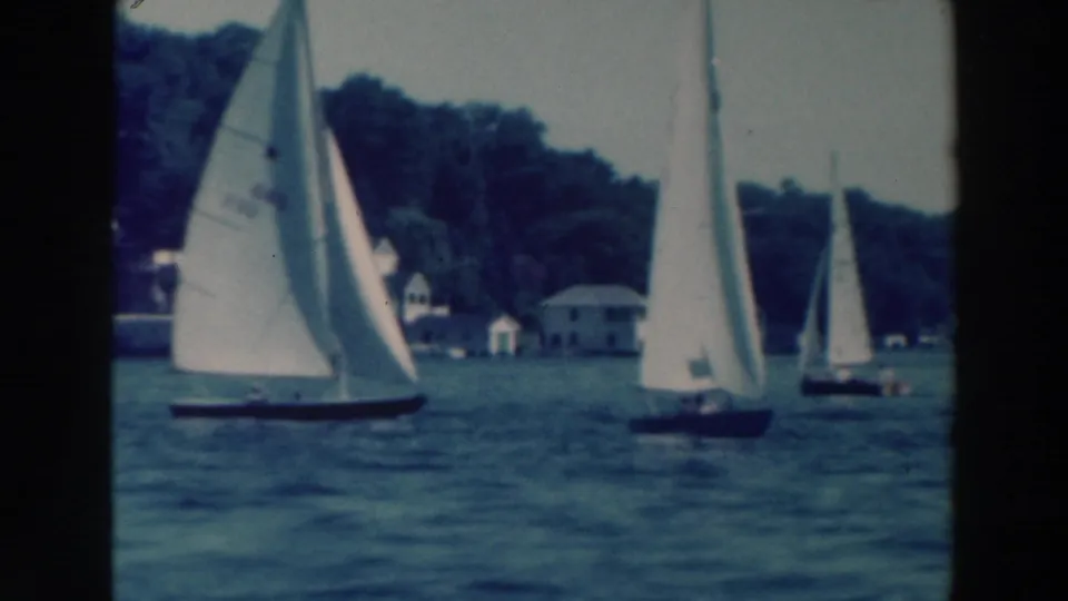1964 Wisconsin home-movie frame of sailboats on calm water, with small shore buildings beyond them.