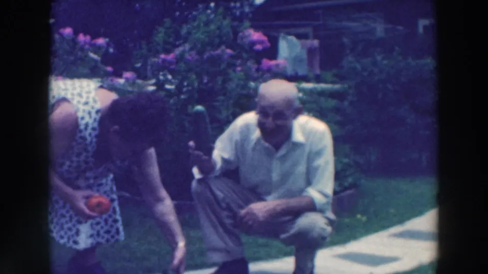 1964 Tustin, Wisconsin 8mm frame showing the couple continuing to arrange garden vegetables in a backyard lawn setting.
