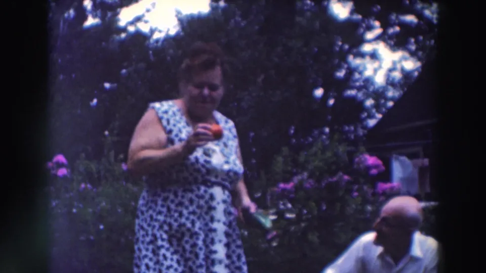 In the backyard garden, the woman looks down at a red tomato as the squatting man remains partly visible nearby.