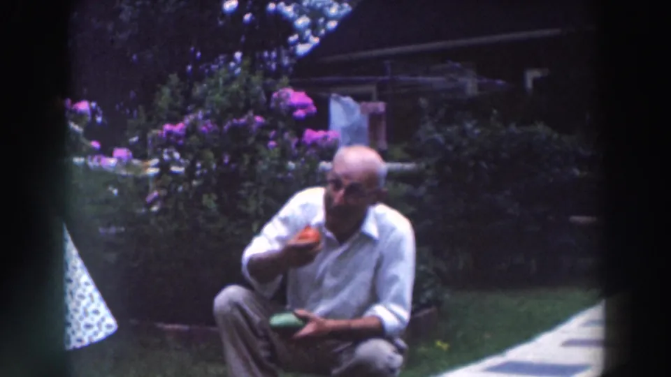 In the 1964 home-movie frame, the crouching man holds produce in a garden with flowers, lawn, and a walkway behind him.