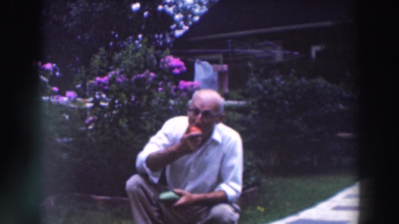 Older man with glasses crouches in a garden while eating a red tomato.