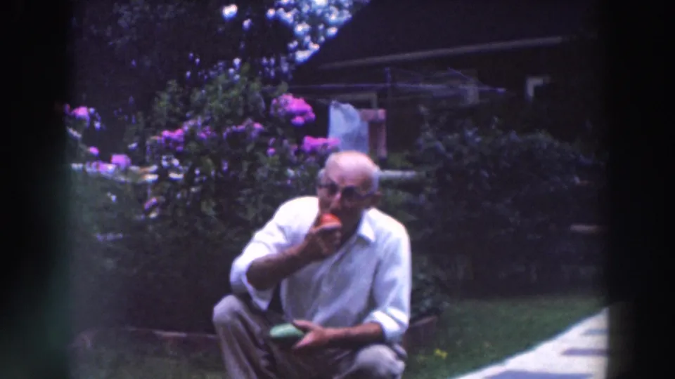 In 1964 Tustin, Wisconsin home-movie footage, an older man in a white shirt crouches in a garden and bites a tomato.