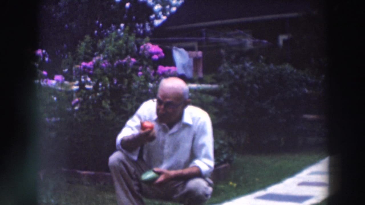 The older man remains crouched in the Tustin garden, holding a red tomato near his mouth with flowers and laundry behind him.