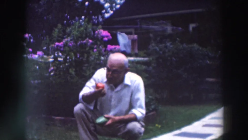 The older man remains crouched in the Tustin garden, holding a red tomato near his mouth with flowers and laundry behind him.