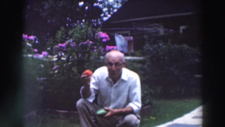 An older man in glasses faces the camera in the garden, holding a tomato in one hand and a green vegetable in the other.