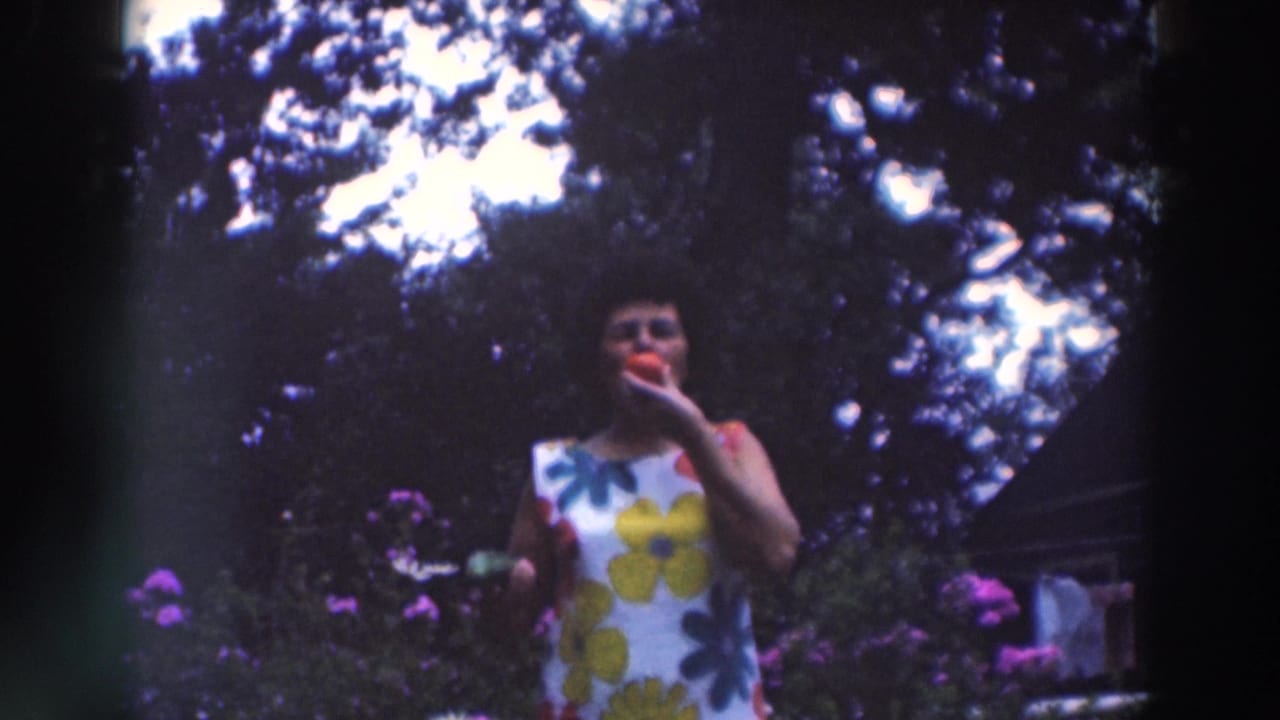 Adult woman in a sleeveless white floral dress holds a red apple near her mouth in the 1964 garden footage.