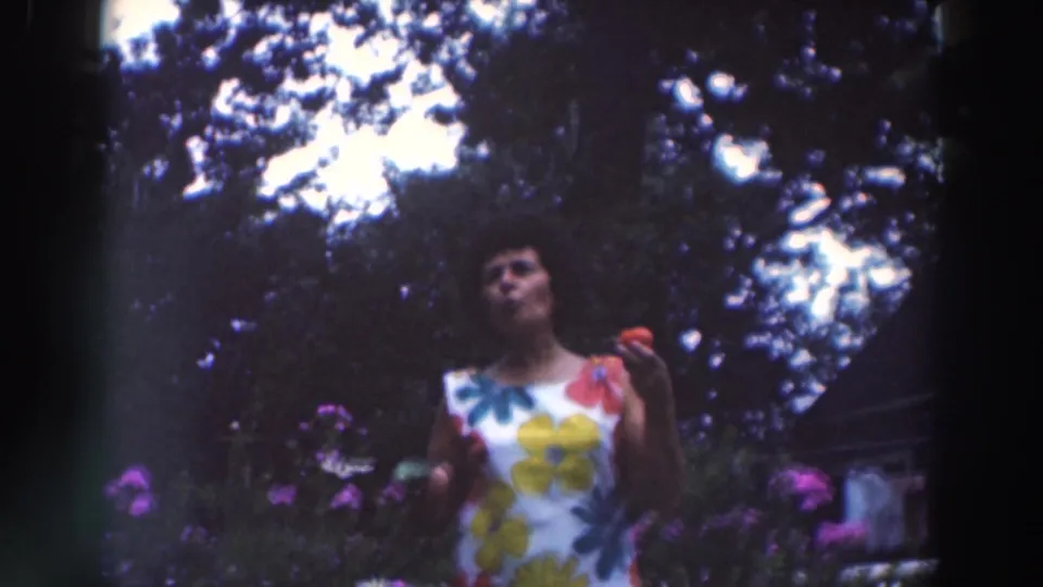 Adult woman appears to chew after biting a red apple, framed by garden flowers and trees in 1964 Tustin footage.