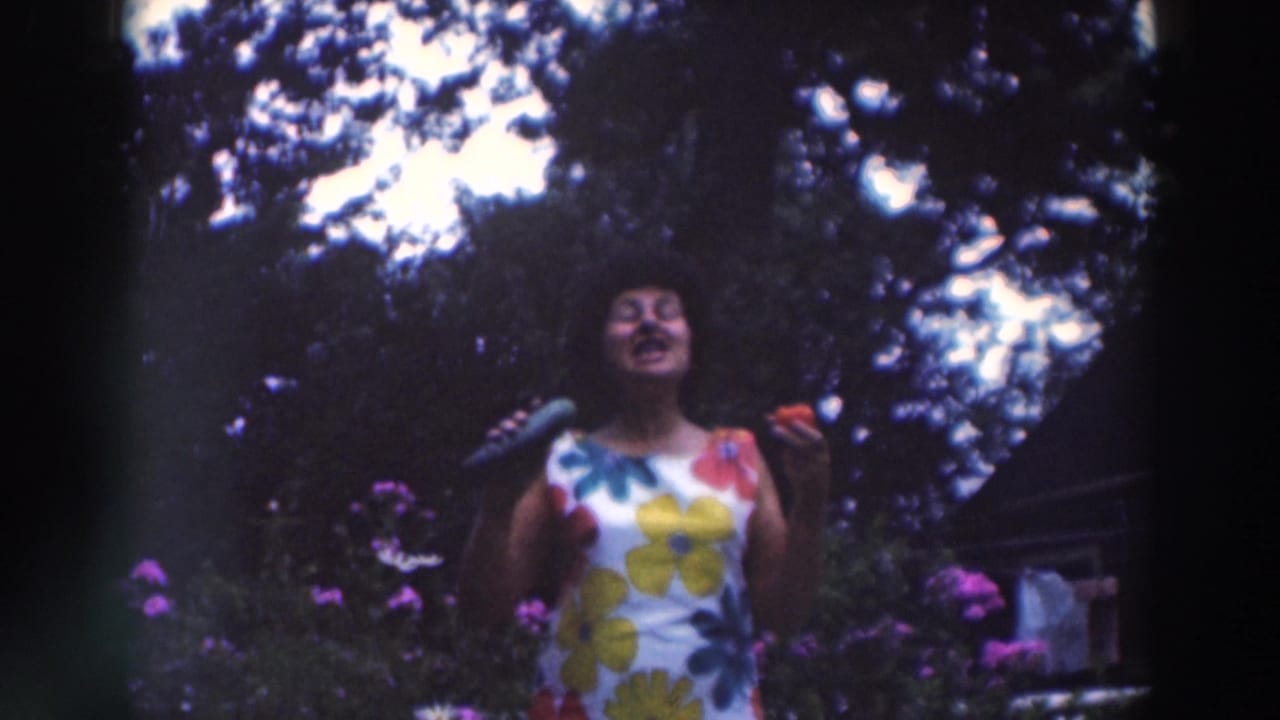 The woman appears animated in the 1964 Tustin garden scene, holding a cucumber and tomato near tall flowers and trees.