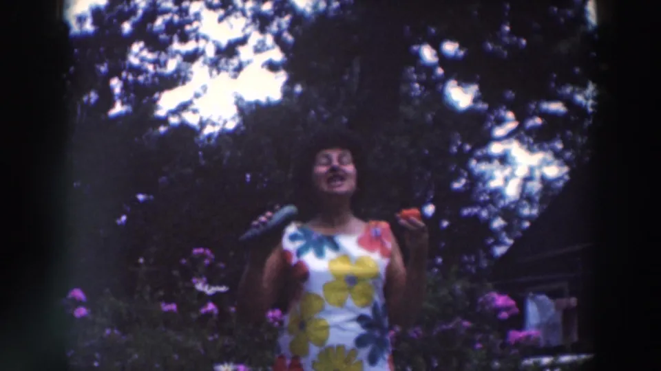 The woman appears animated in the 1964 Tustin garden scene, holding a cucumber and tomato near tall flowers and trees.