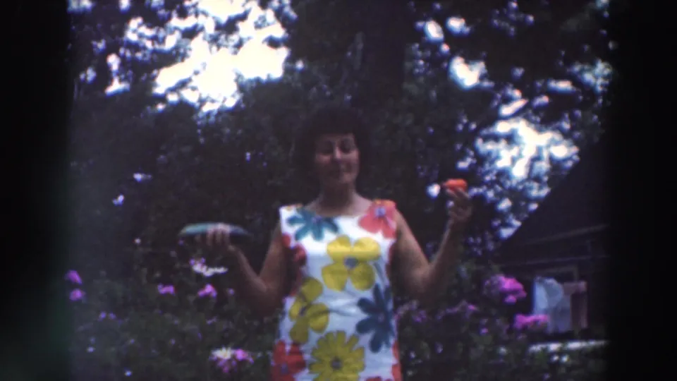 A woman in a bold floral dress gestures in a Wisconsin garden, with purple flowers and tree shadows around her.