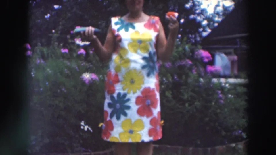 A fuller 1964 view shows the woman in her white flower dress holding a cucumber and tomato amid garden plants and trees.