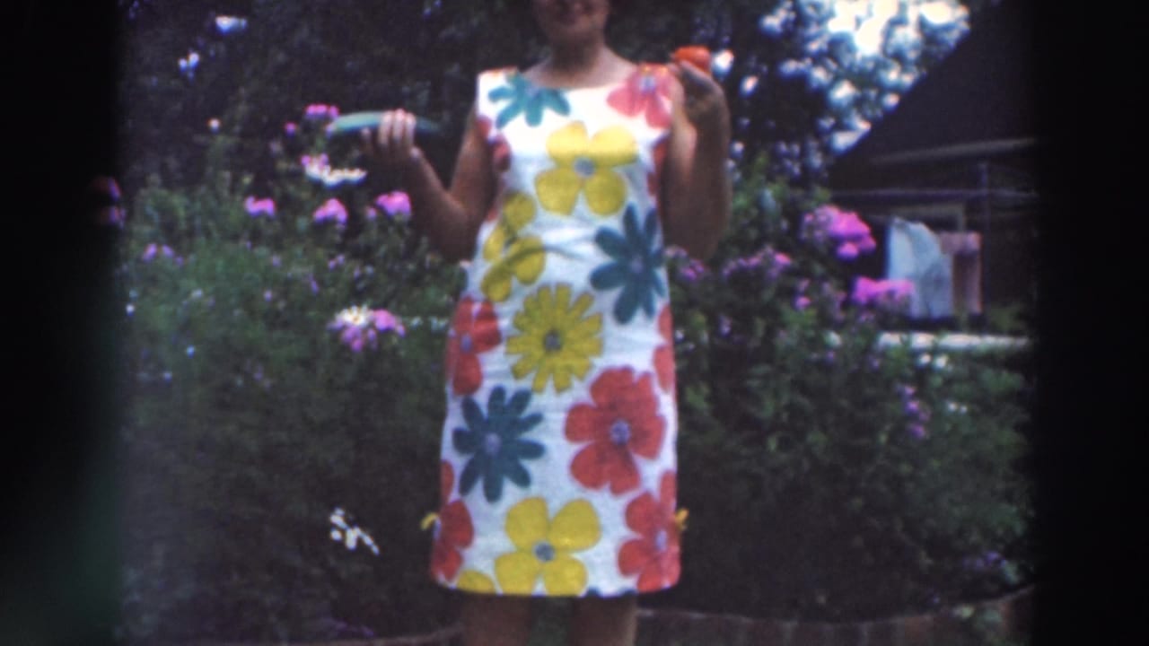 Woman in a bright floral dress standing in a garden holding a red treat and green object.