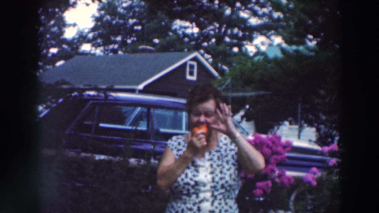 Woman in a patterned dress bites a red tomato in a backyard near flowering shrubs and a parked car.