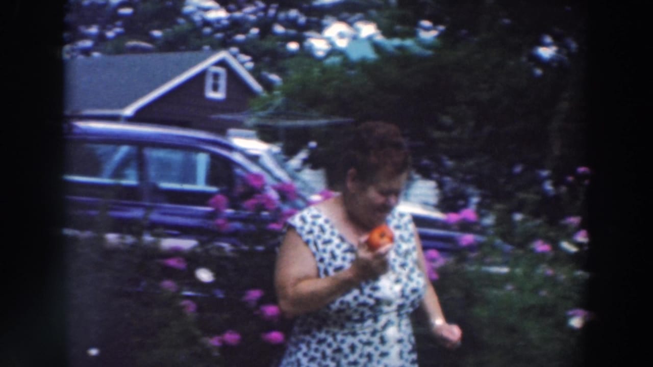 Woman looks down while holding a tomato beside purple flowers, lawn, and a parked car.