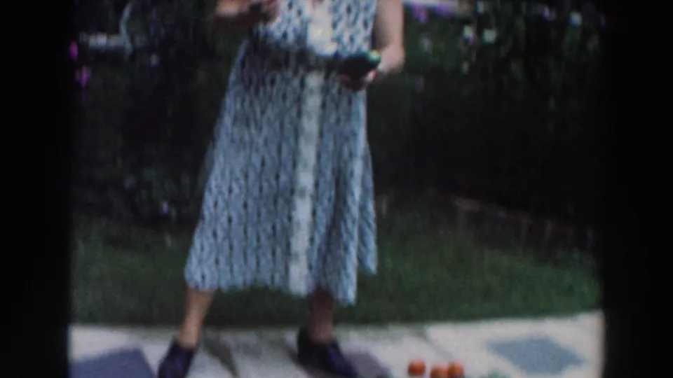 A partial 1964 frame shows the woman standing on grass beside tomatoes and cucumbers set on the walkway in a backyard garden area.