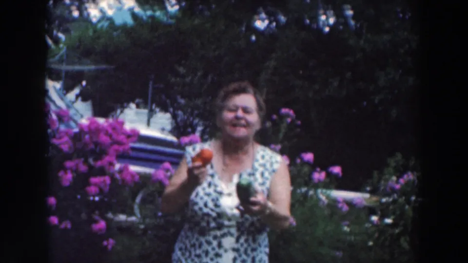 In metadata-supported Tustin, Wisconsin, 1964, an adult woman smiles in a flower garden while holding a tomato and green pepper.
