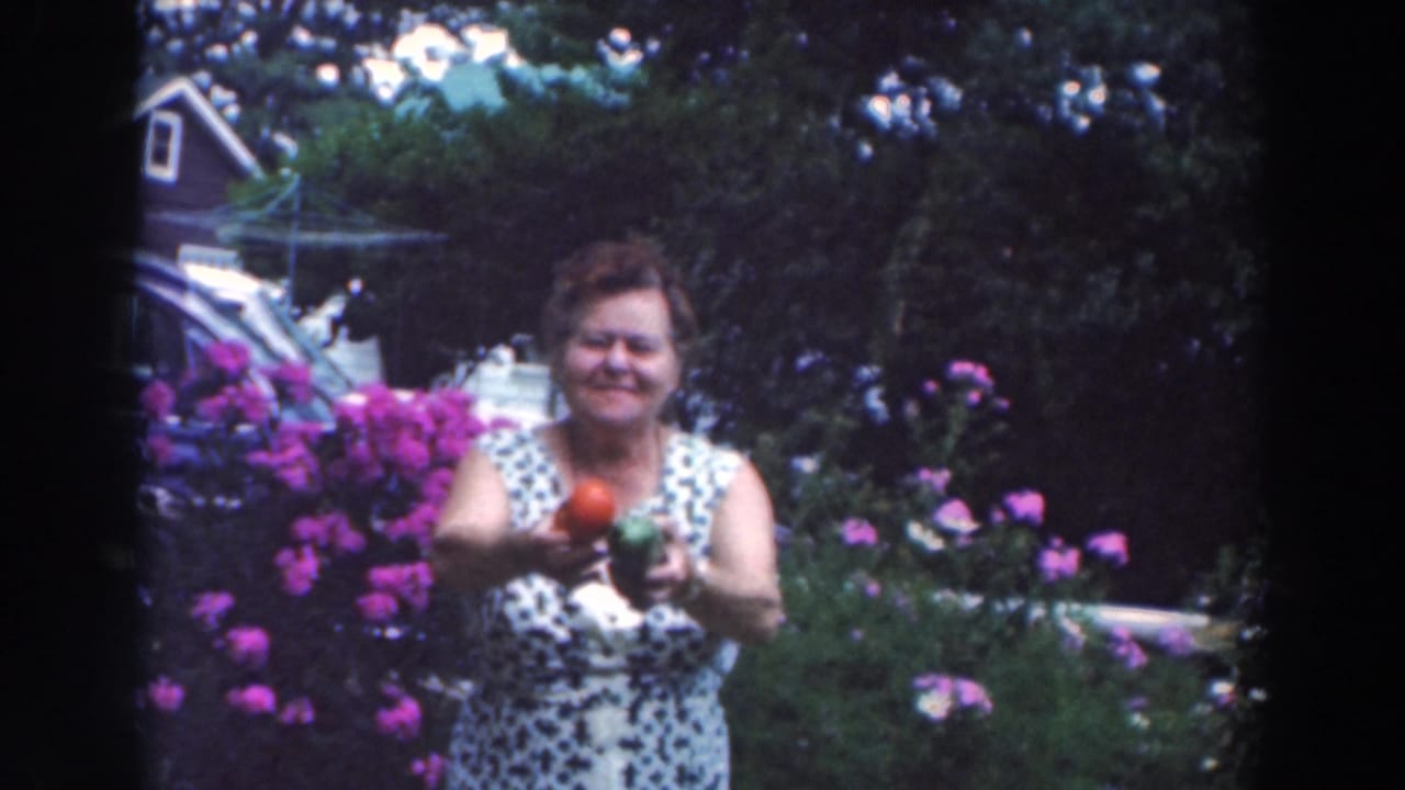 Woman smiles toward the camera while extending a red tomato and green pepper in a garden.