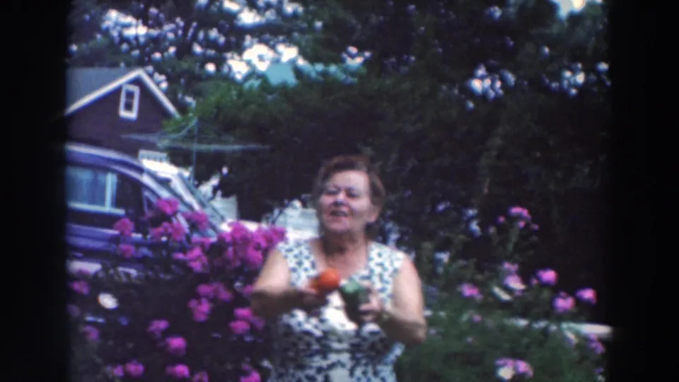 The woman remains in the garden with tomato and pepper visible, surrounded by pink flowers, greenery, and nearby residential yard features.