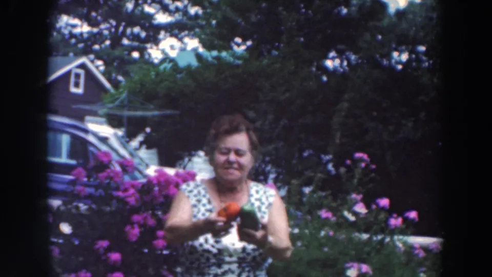 Near the end of the 1964 8mm clip, the woman looks down at the produce while standing among flowers and dense garden foliage.