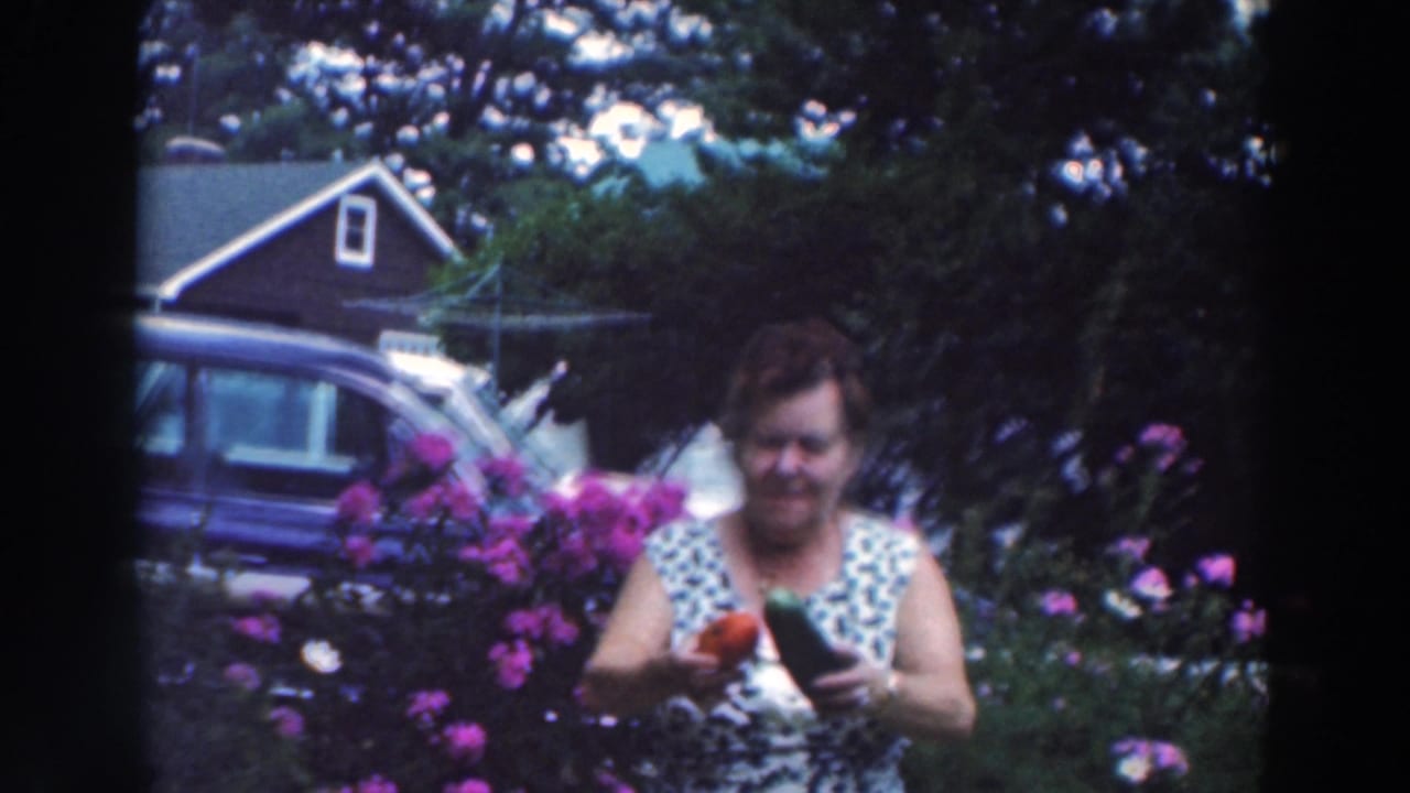 Woman in a flower garden holding a red pepper and cucumber near a parked car and house.