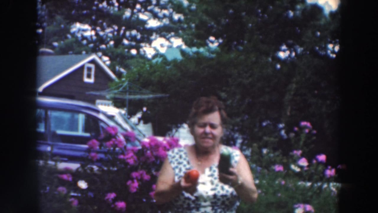In the Tustin, Wisconsin home-movie frame, a woman faces the camera holding a red pepper and cucumber in a blooming residential garden.