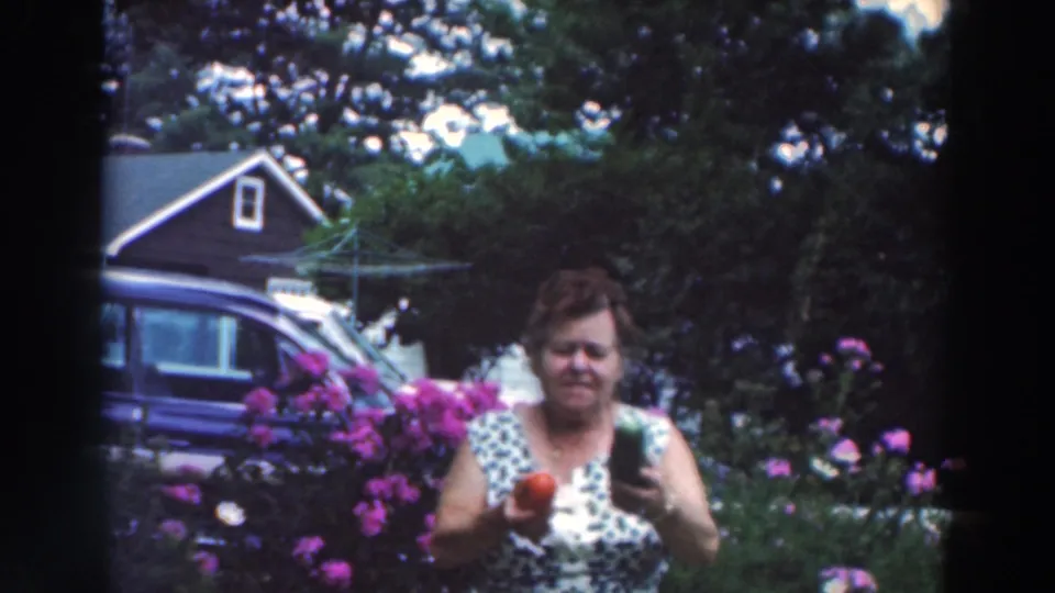In the Tustin, Wisconsin home-movie frame, a woman faces the camera holding a red pepper and cucumber in a blooming residential garden.