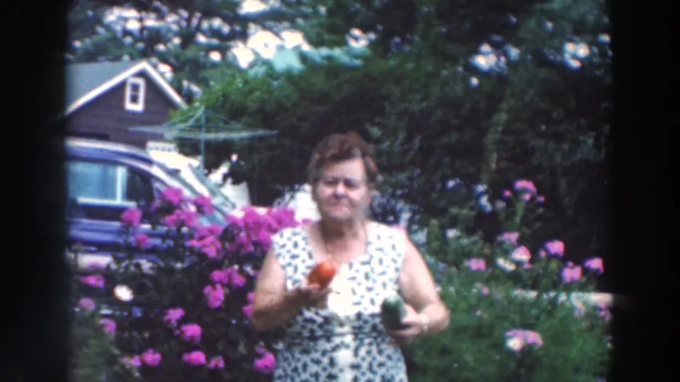 A woman in a patterned sleeveless dress stands in a 1964 Wisconsin garden holding a red pepper, surrounded by flowers and residential details.