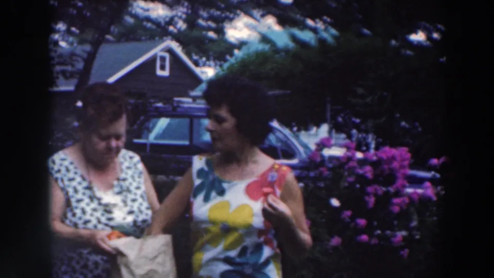 Two adult women in summer dresses stand together in the Tustin, Wisconsin garden, with one holding a paper bag near flowers and a parked car.