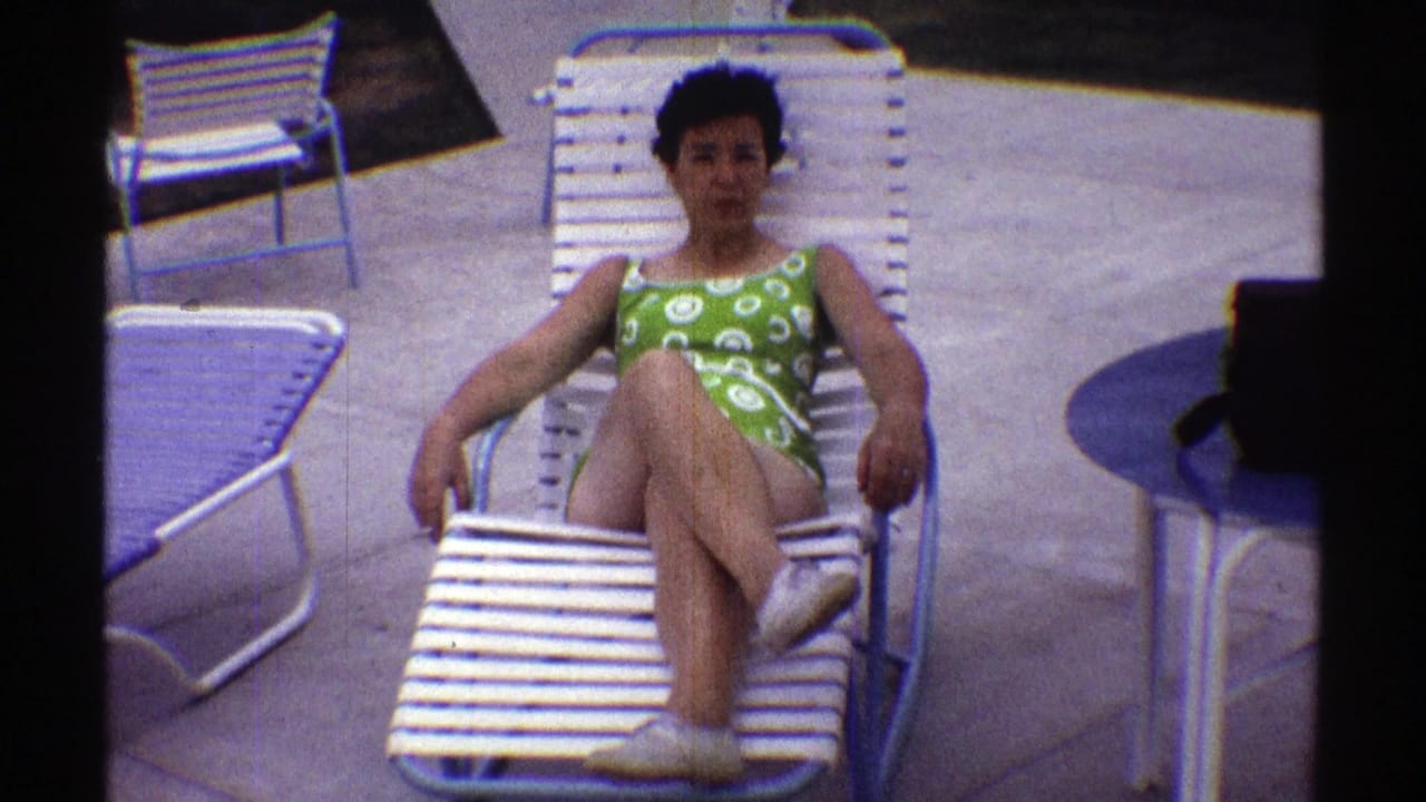 Nebraska 1969 frame showing a woman in a green swimsuit seated on a white poolside lounge chair, with blue table and patio chairs around her.
