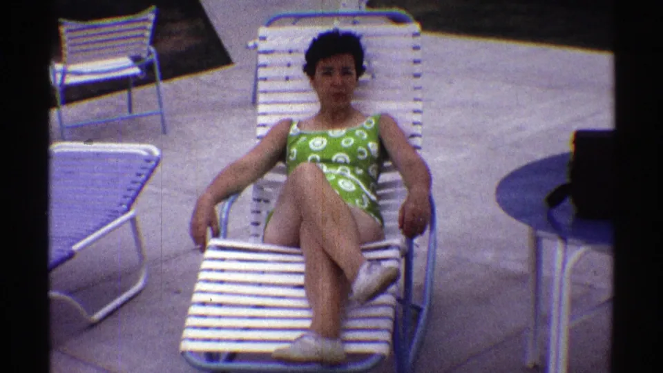 Nebraska 1969 frame showing a woman in a green swimsuit seated on a white poolside lounge chair, with blue table and patio chairs around her.