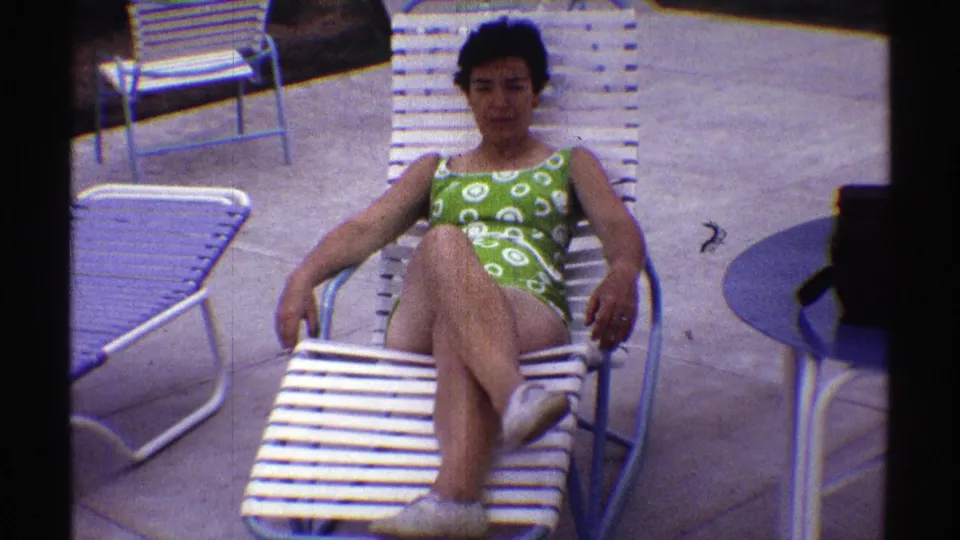 A 1969 Nebraska home-movie still shows the woman reclining on a white slatted lounge chair, framed by nearby poolside furniture.