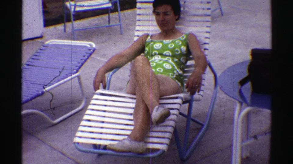 Final frame of the Nebraska 1969 poolside clip, with the woman angled on a lounge chair and nearby chairs and a blue table visible.