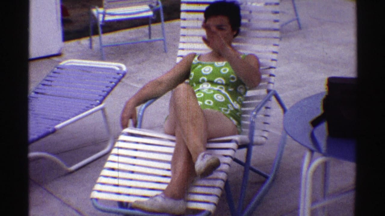 In Nebraska 1969 home-movie footage, a woman in a green patterned one-piece swimsuit sits on a white slatted lounge chair.