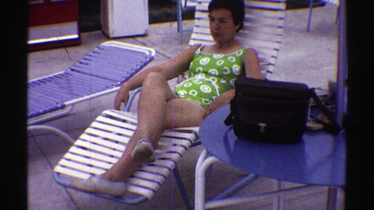 In 1969 Nebraska home-movie footage, a woman reclines on a white and blue striped lounge chair near a blue table and black handbag.