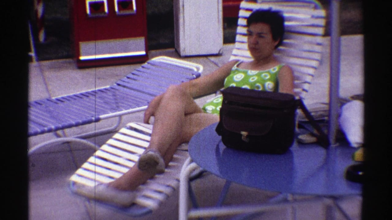 In Nebraska 1969 home-movie footage, a woman in green patterned swimwear reclines on a white poolside lounge chair near a blue table and black handbag.
