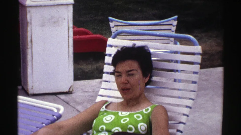 Nebraska 1969 frame of a woman seated on a white lounge chair, with red outdoor furniture and poolside paving visible behind her.