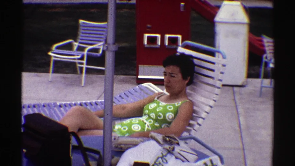 Wide 1969 Nebraska poolside view with the woman reclining, a blue table and black handbag nearby, white lounge chairs, and a red vending machine in back.