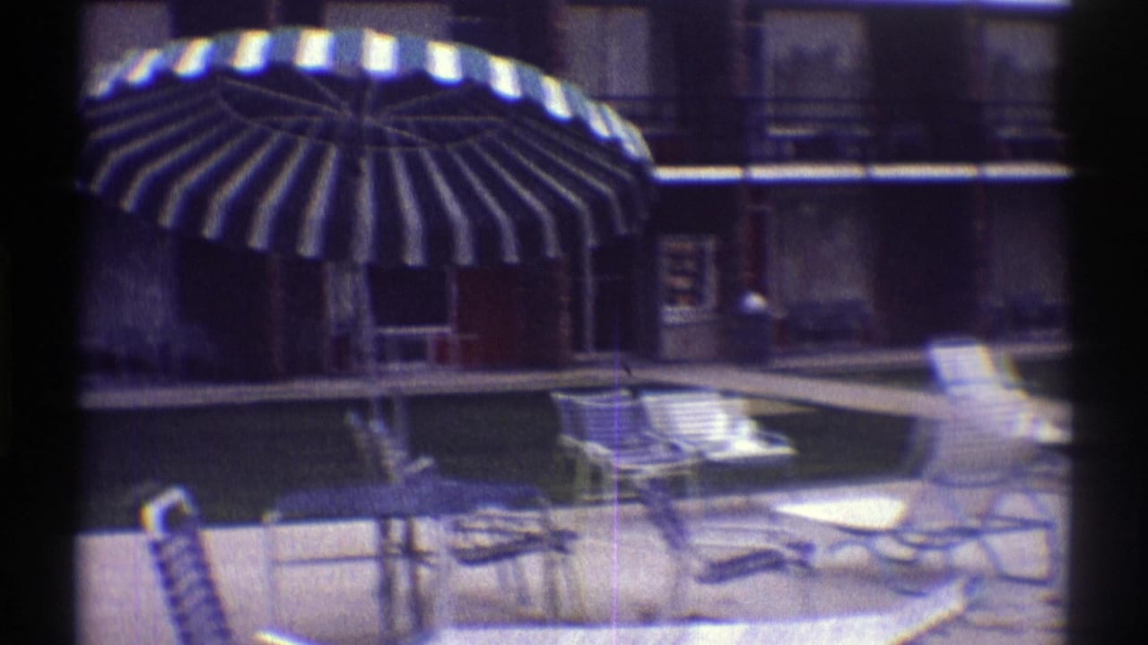 1969 Nebraska motel courtyard with a swimming pool, striped umbrella, metal lounge chairs, and brick unit buildings in soft 8mm color.