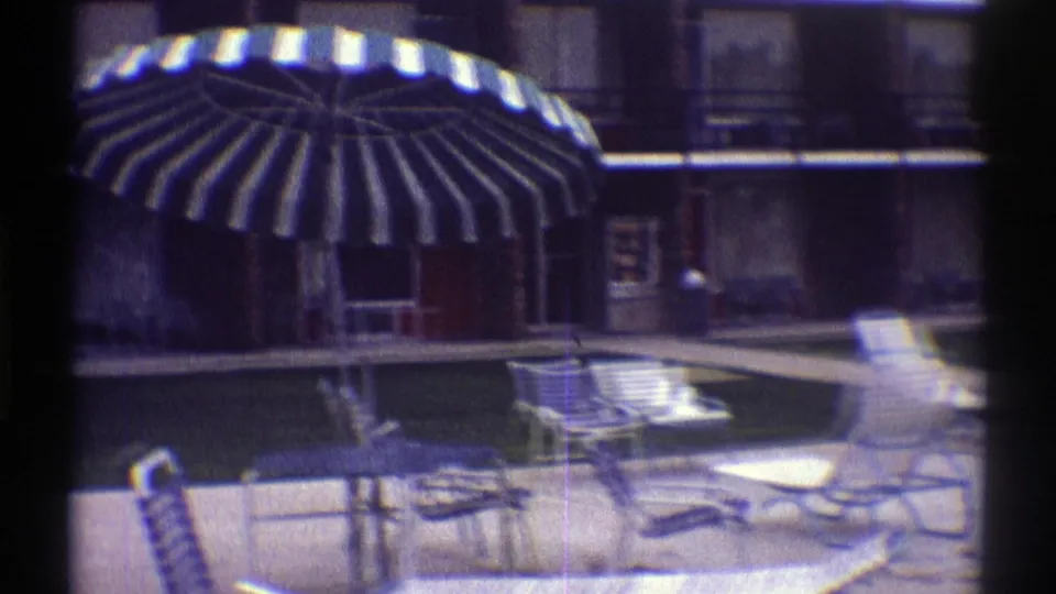 1969 Nebraska motel courtyard with a swimming pool, striped umbrella, metal lounge chairs, and brick unit buildings in soft 8mm color.