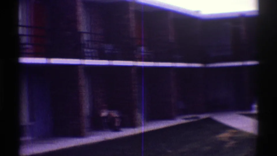 Dim 8mm view of the Mos Motel courtyard area, showing balcony railings, brick walls, a walkway, and a small seated person.
